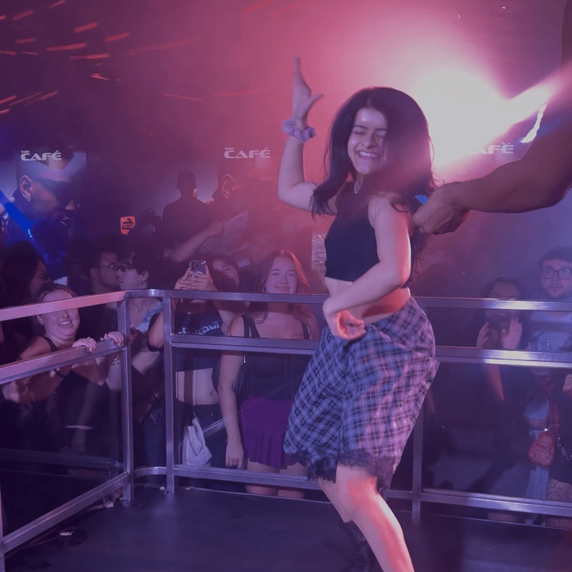 Nush dancing and laughing on a stage under pink club lights while the crowd cheers